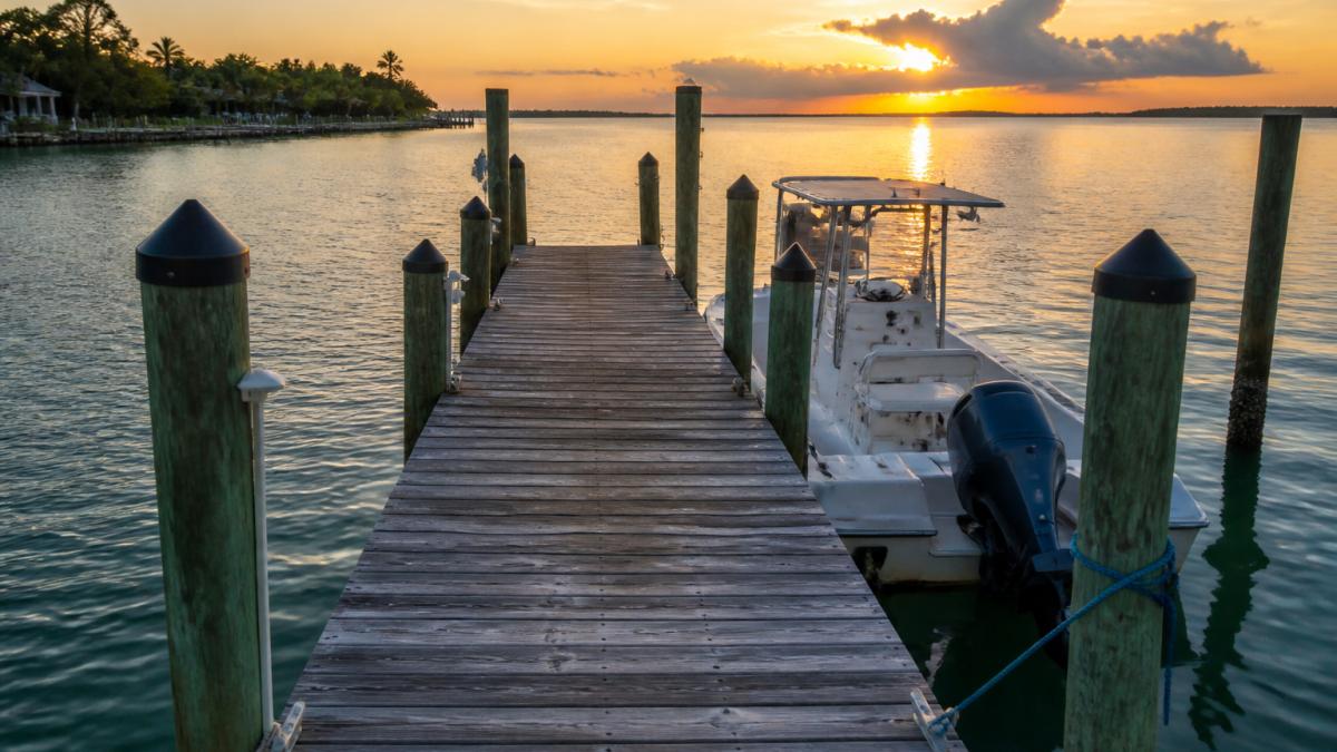A weathered wooden dock extending into calm coastal water at golden hour, with pilings visible and a small boat moored alongside, showing typical saltwater exposure conditions
